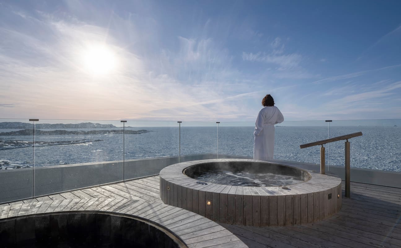 Person in white robe on a wooden deck with steaming hot tubs overlooking an ocean with ice and land