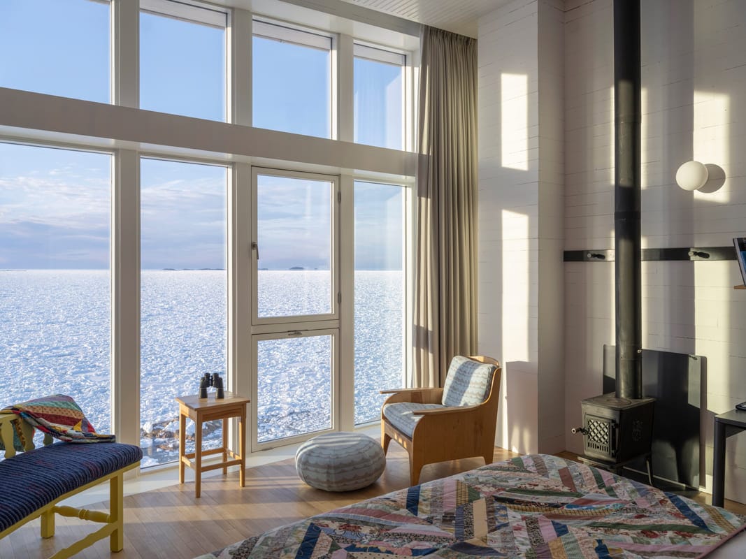 Modern room with large windows facing a frozen sea, featuring an armchair, wooden bench, stove, and colorful quilt